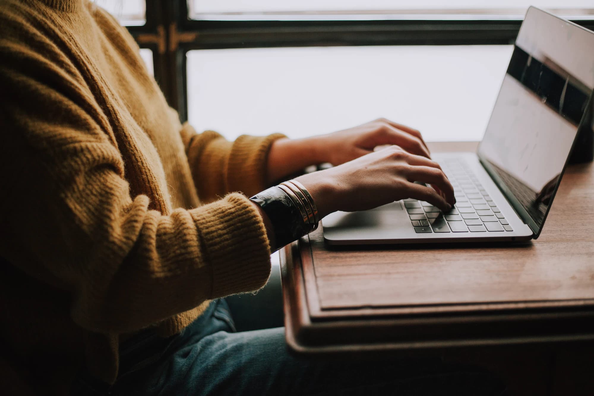 Person typing on a laptop at a wooden desk by a window