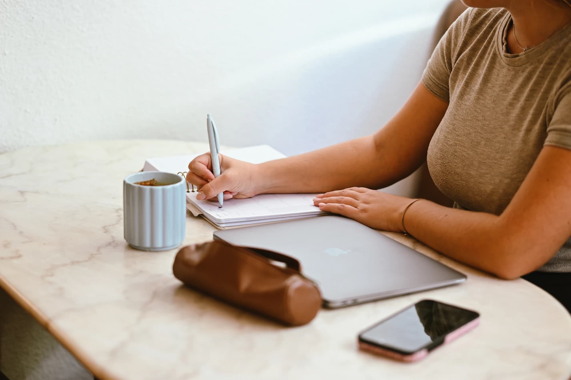 Person writing in a planner at a marble café table beside a laptop, phone and mug