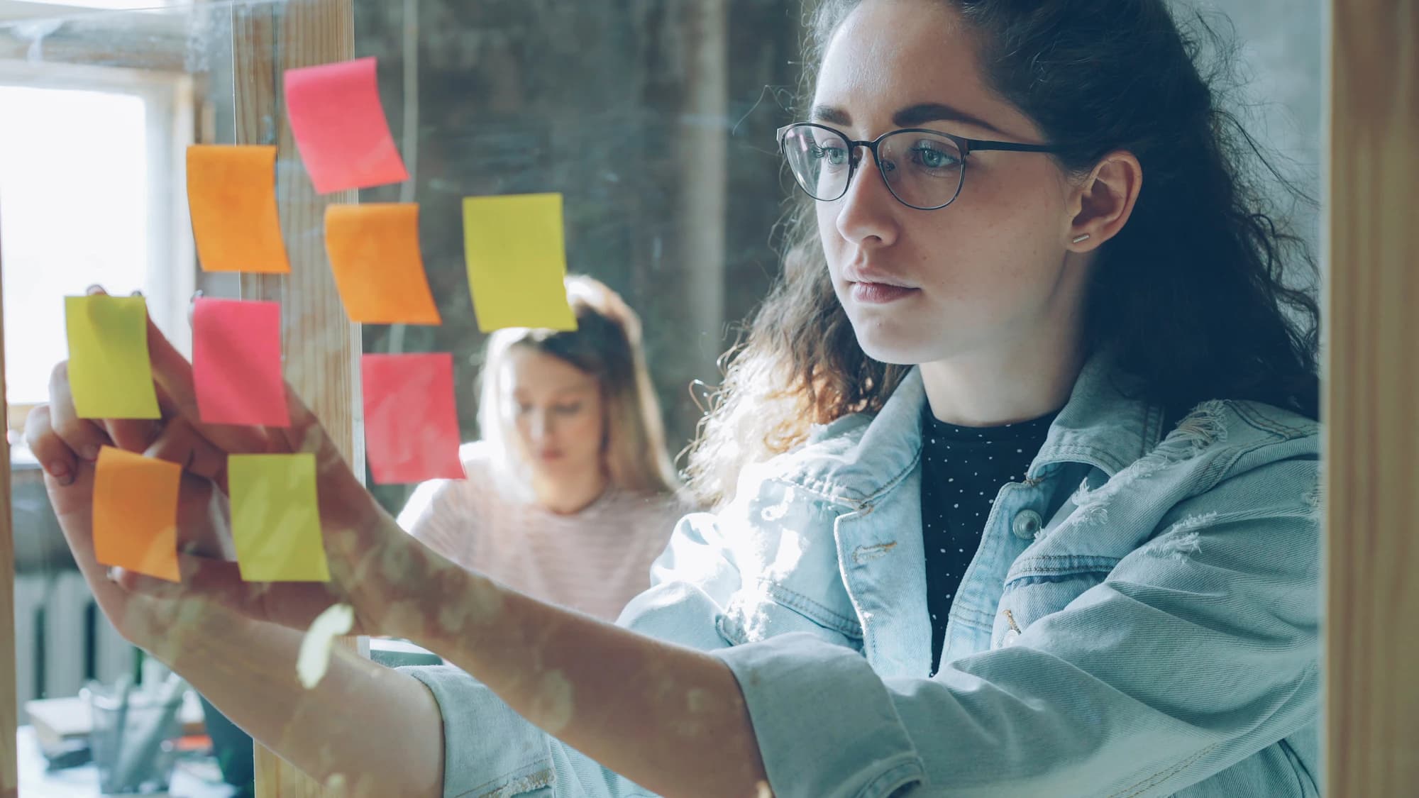 Woman arranging colourful sticky notes on a glass wall during a planning session, a colleague working behind her