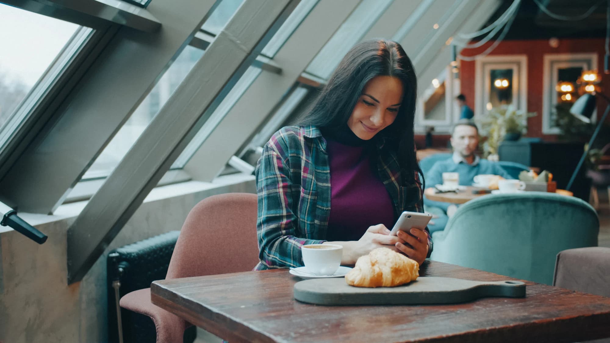 Woman smiling at her phone at a wooden café table with coffee and a croissant