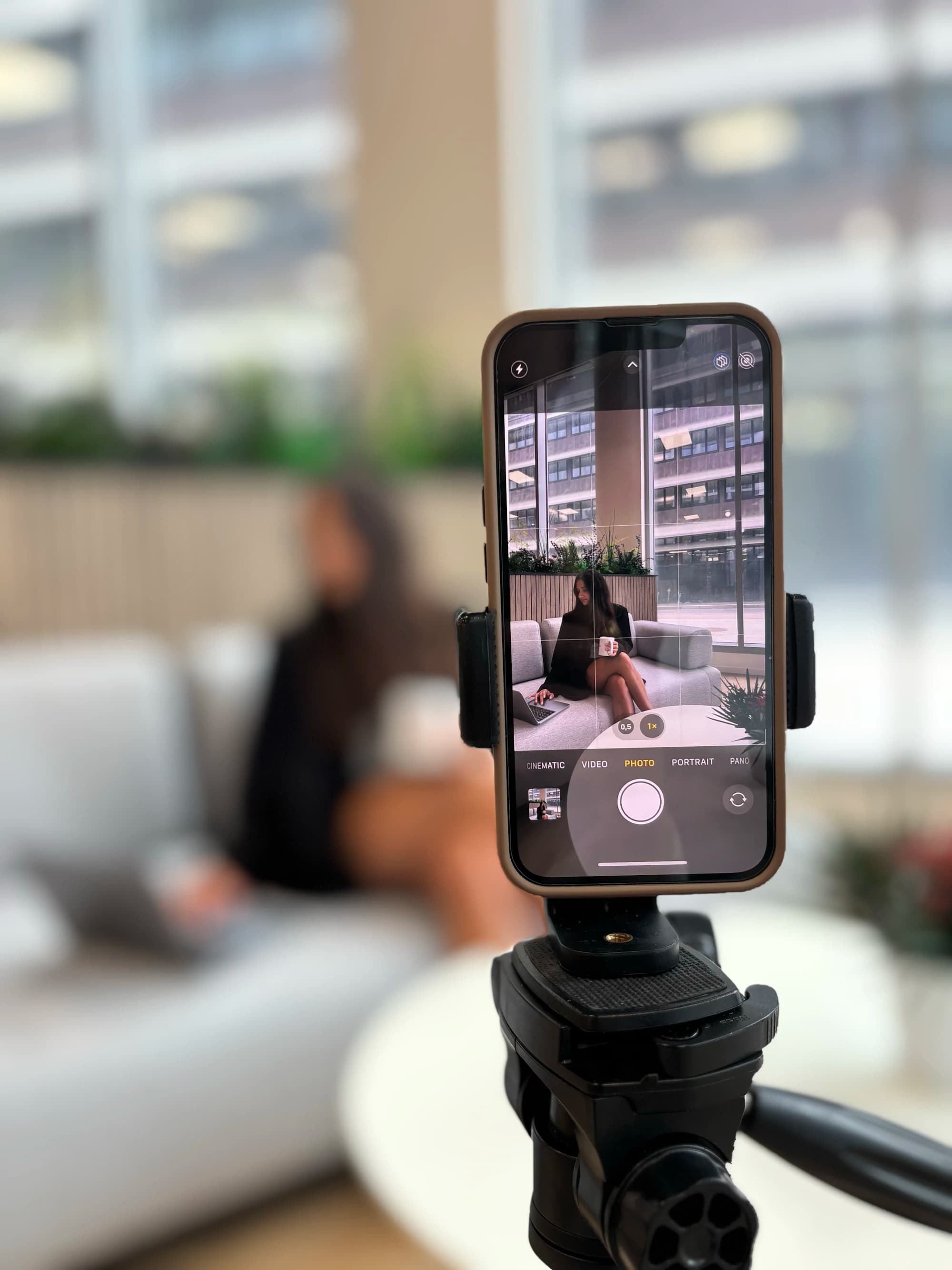 Smartphone on a tripod framing a woman working on a sofa in a bright, plant-filled office