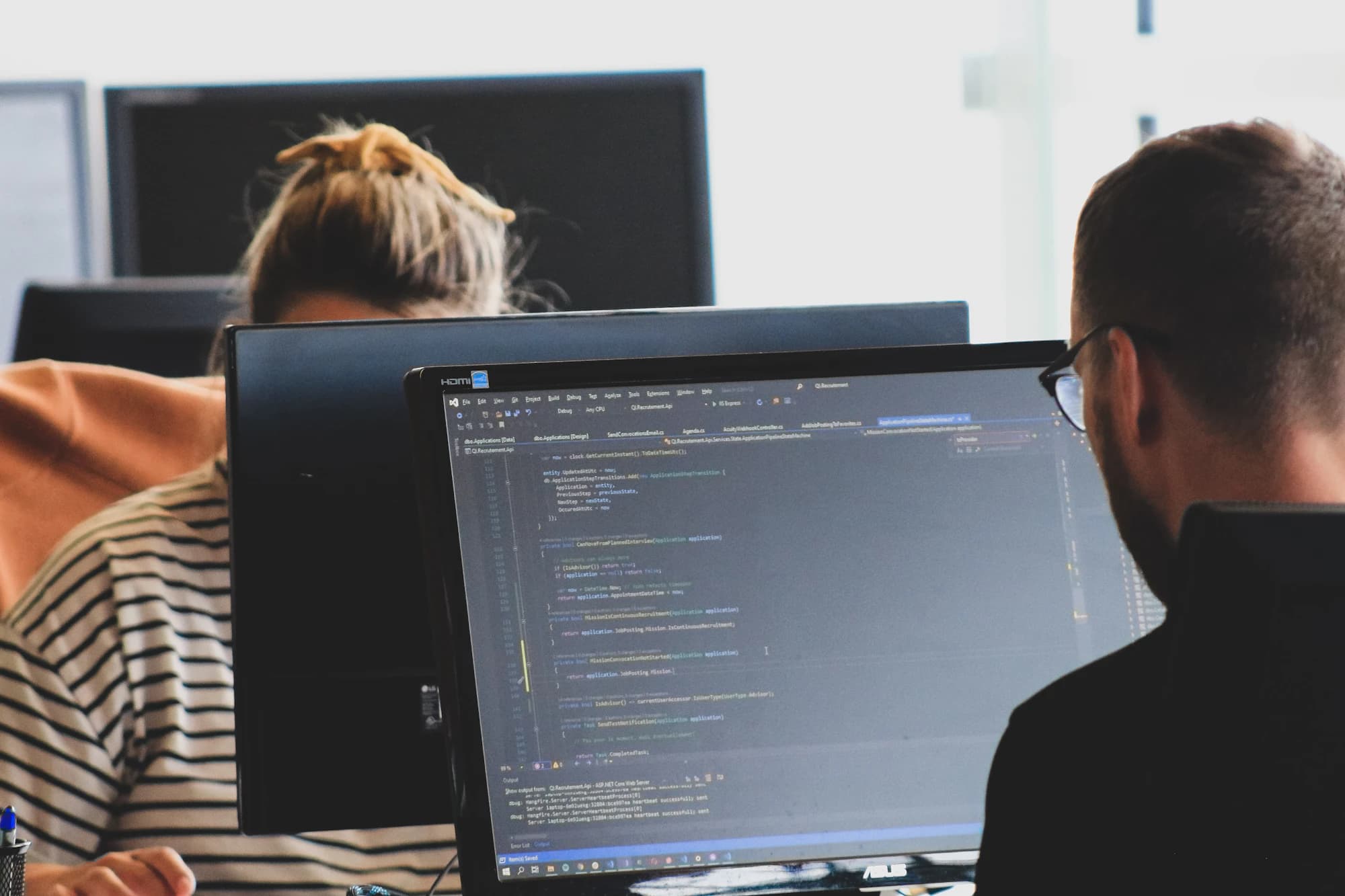 Two developers working at monitors in a bright office, code open in an editor on the nearest screen